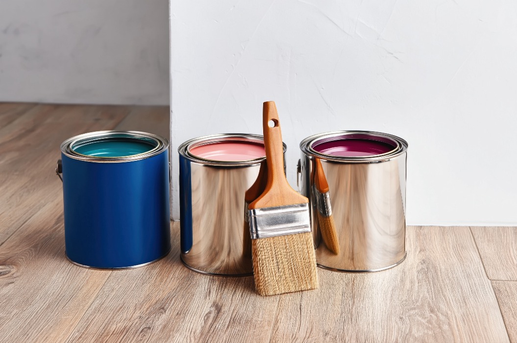 Geöffnete Farbdosen und ein Pinsel liegen auf einem Holzboden in der Nähe einer weißen Wand, bereit für die Renovierung des Hauses. Eine Szene aus dem Bereich der Innenraumgestaltung, in deren Mittelpunkt das Streichen und die Dekoration der Wände stehen