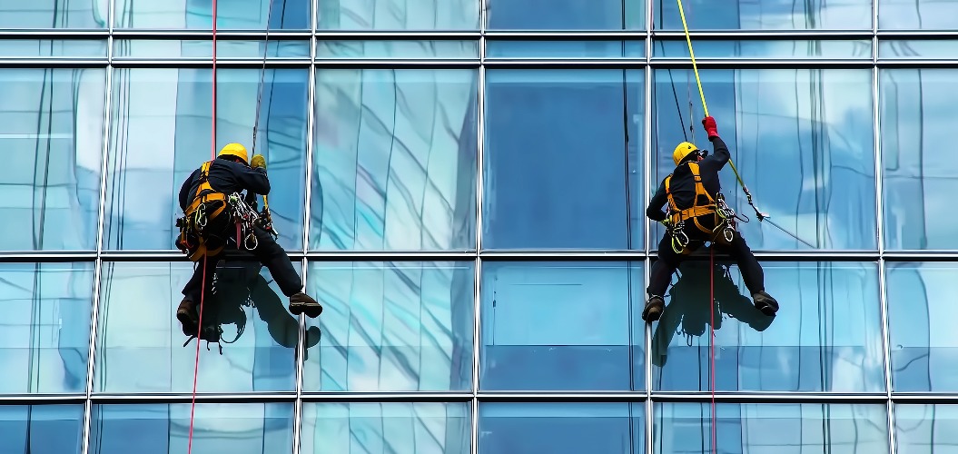 Zwei Arbeiter mit Schutzhelm und Sicherheitsgurt, die in der Luft schweben und die Außenfassade eines Firmengebäudes oder Wolkenkratzers reinigen